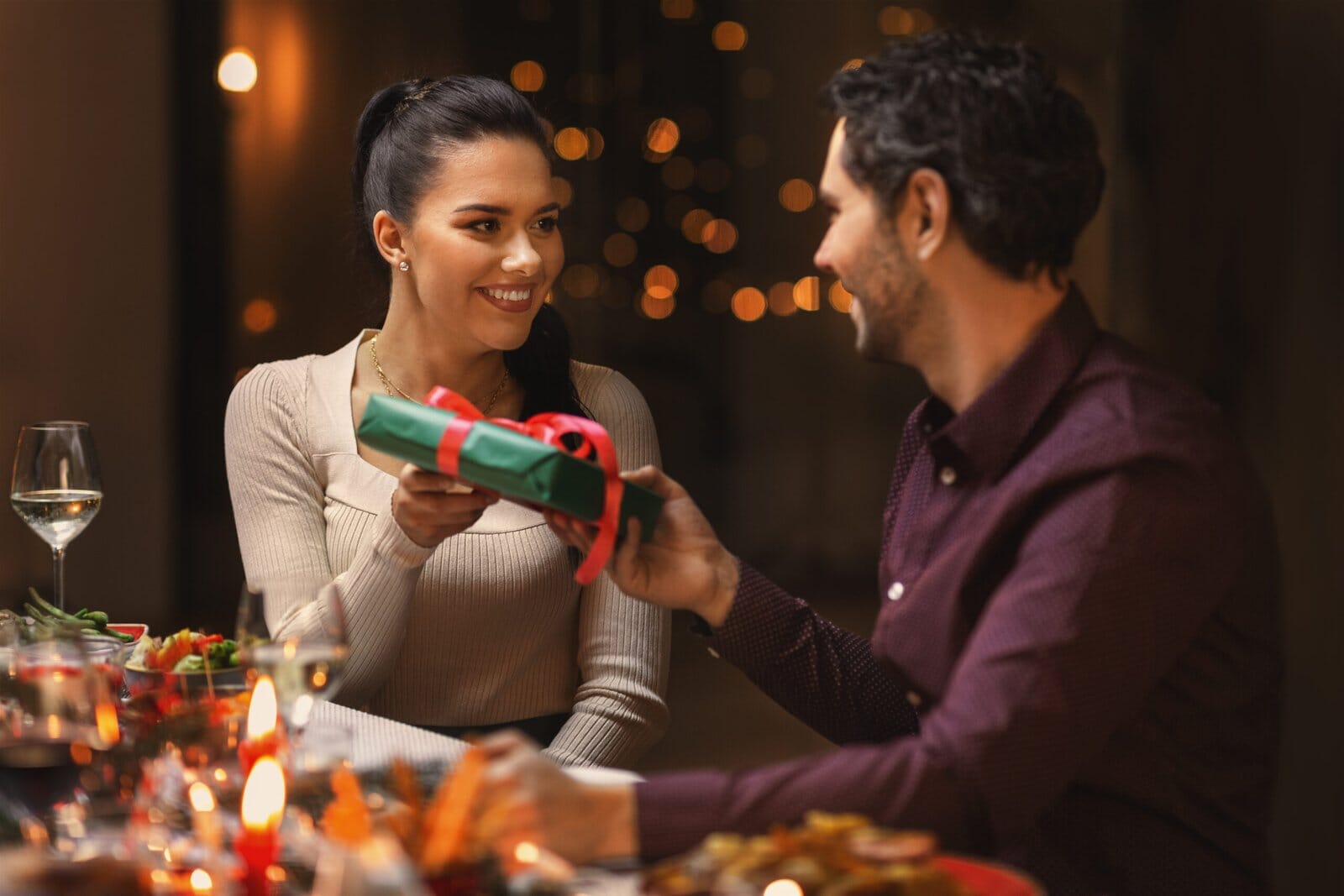 A Woman And A Man Share A Warm Moment As She Hands Him A Wrapped Gift Across A Festive, Candle-Lit Dinner Table.