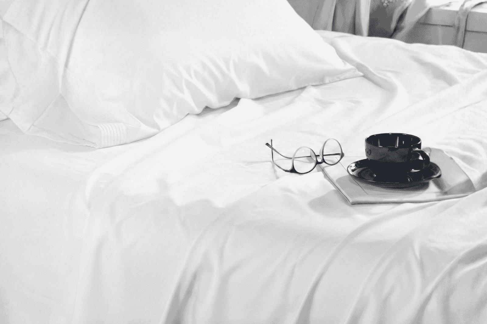 Close-Up Of Crisp White Bed Sheet With A Pair Of Glasses And A Black Cup And Saucer On A Magazine, For A Cozy Minimalist Guest Bedroom.