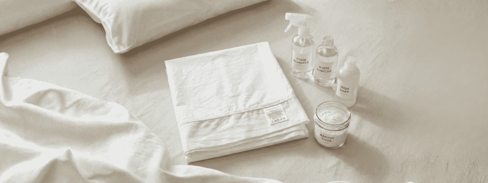 Luxury Sheets Laid On A White Bed Beside Natural Stain-Removal Supplies, Including White Vinegar, Dish Soap, And Baking Soda.